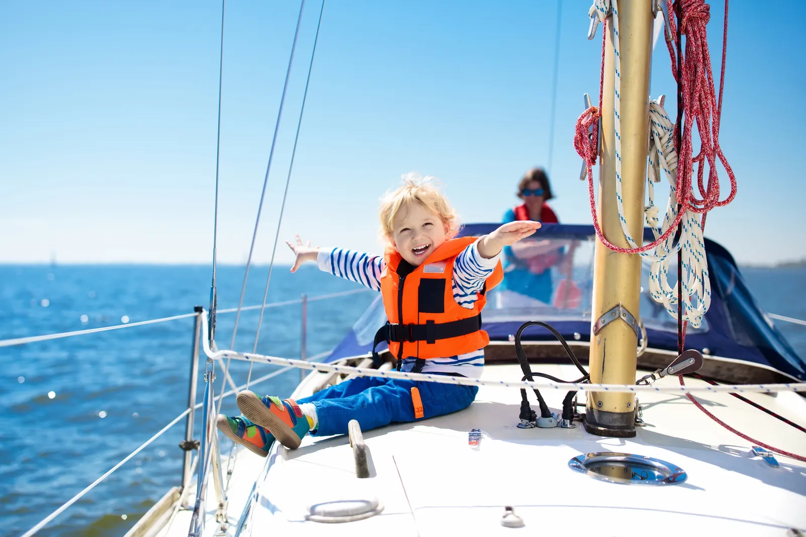 A child in a life jacket grinning on the foredeck of a yacht under sail